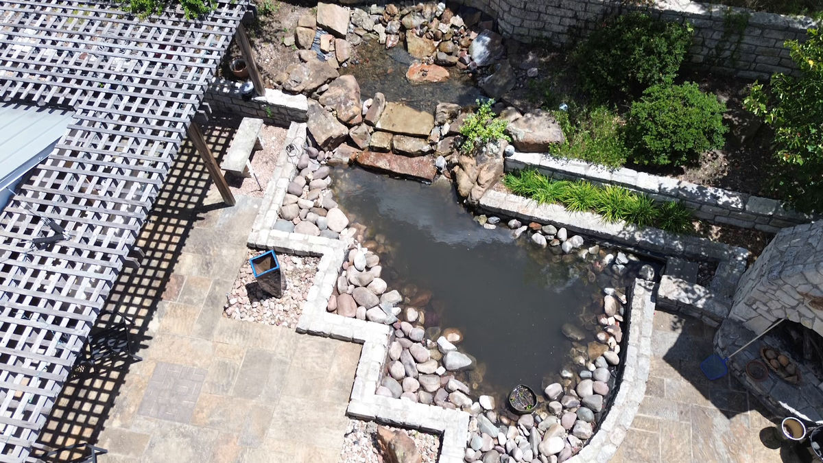 An aerial view of a stone-walled pond with a flagstone terrace, a boulder stream, a lattice pergola and a stone outdoor fireplace.