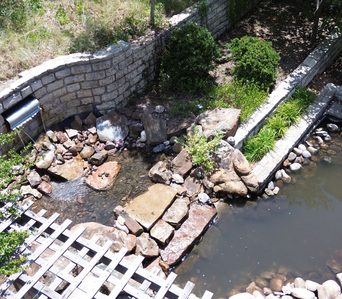 An aerial view of a tall curved limestone-block wall with a metal spillway, a boulder stream running down into a pond, and a stone outdoor fireplace at the far end.
