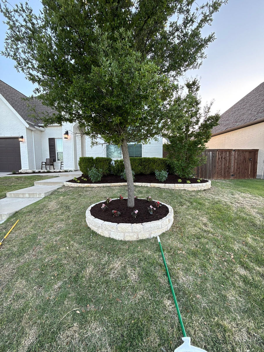 A circular limestone-block tree ring filled with dark mulch and pink begonias, set in the front lawn of the same house.