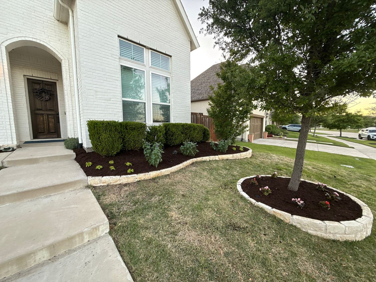 A white brick house at dusk with a stone-edged foundation bed and a separate circular tree bed set in the grass.