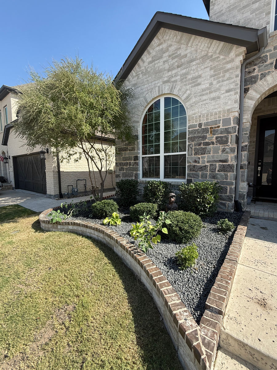 A brick-edged planting bed curving around the corner of the same stone-fronted house, newly planted with shrubs and perennials over gravel mulch.