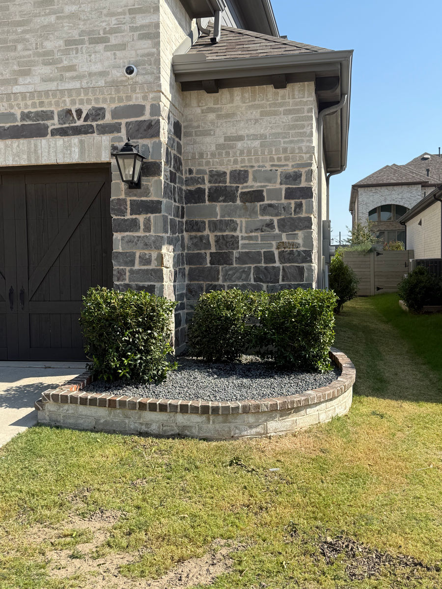 A brick-edged bed of dark gravel mulch and low clipped shrubs curving around the stone corner of the house beside the garage door.