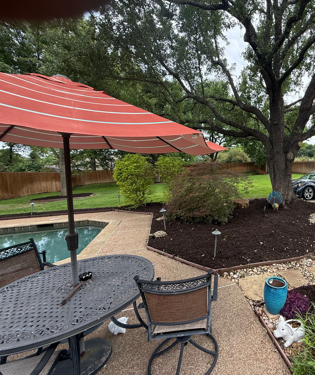 A shaded patio with a table and parasol beside the pool, and a large mulched bed with stone edging cut in beneath a mature oak.