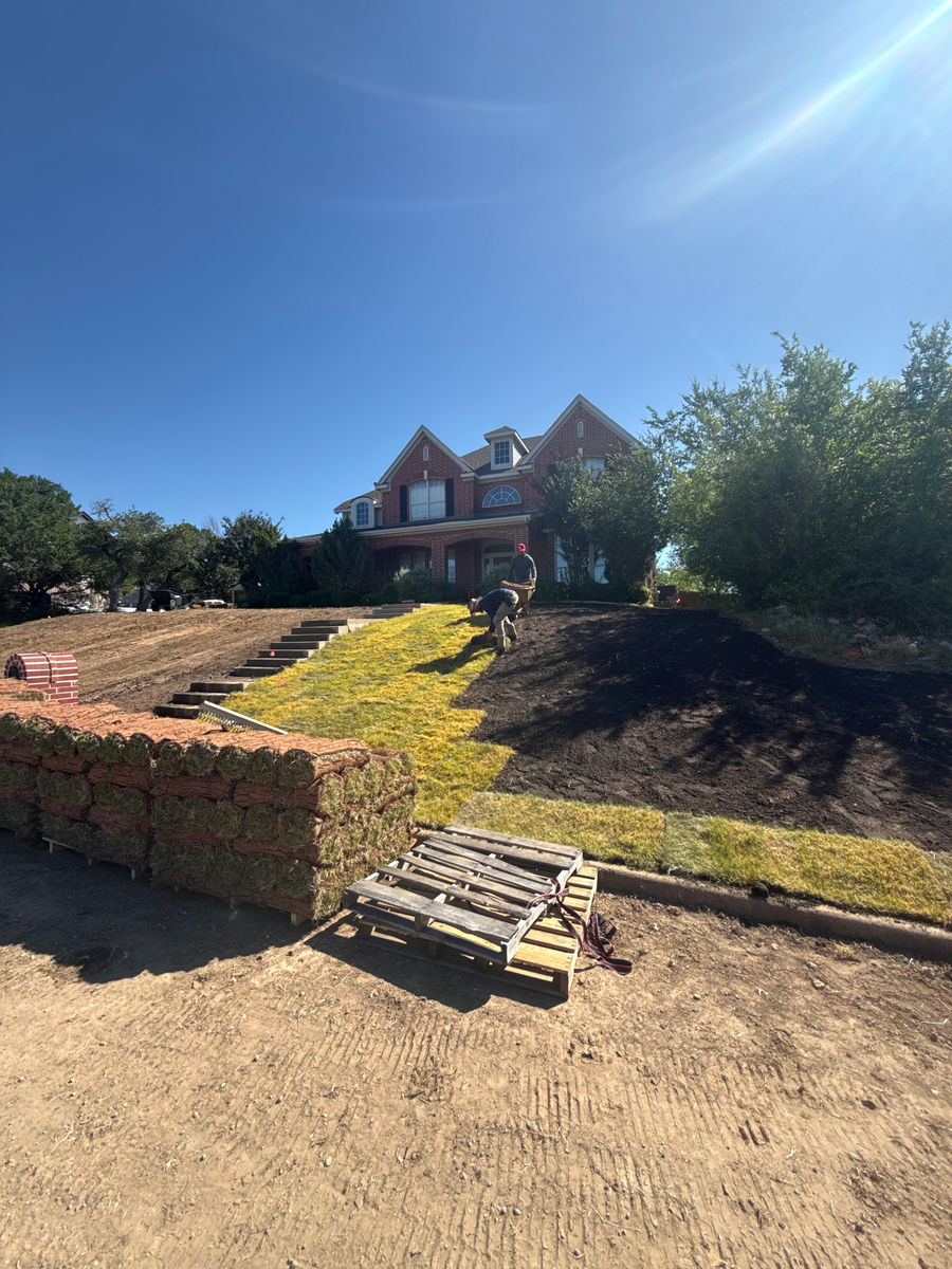 A new limestone retaining wall going in across a bare graded slope, with stacked pallets of block still waiting, below a large brick house.