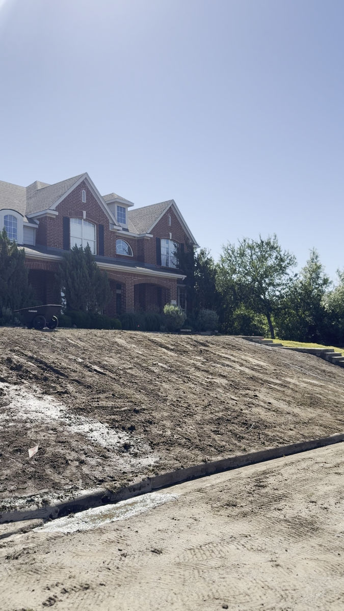 The same front slope graded down to bare soil, with a fresh concrete edge along the drive and stone steps cut into the bank.
